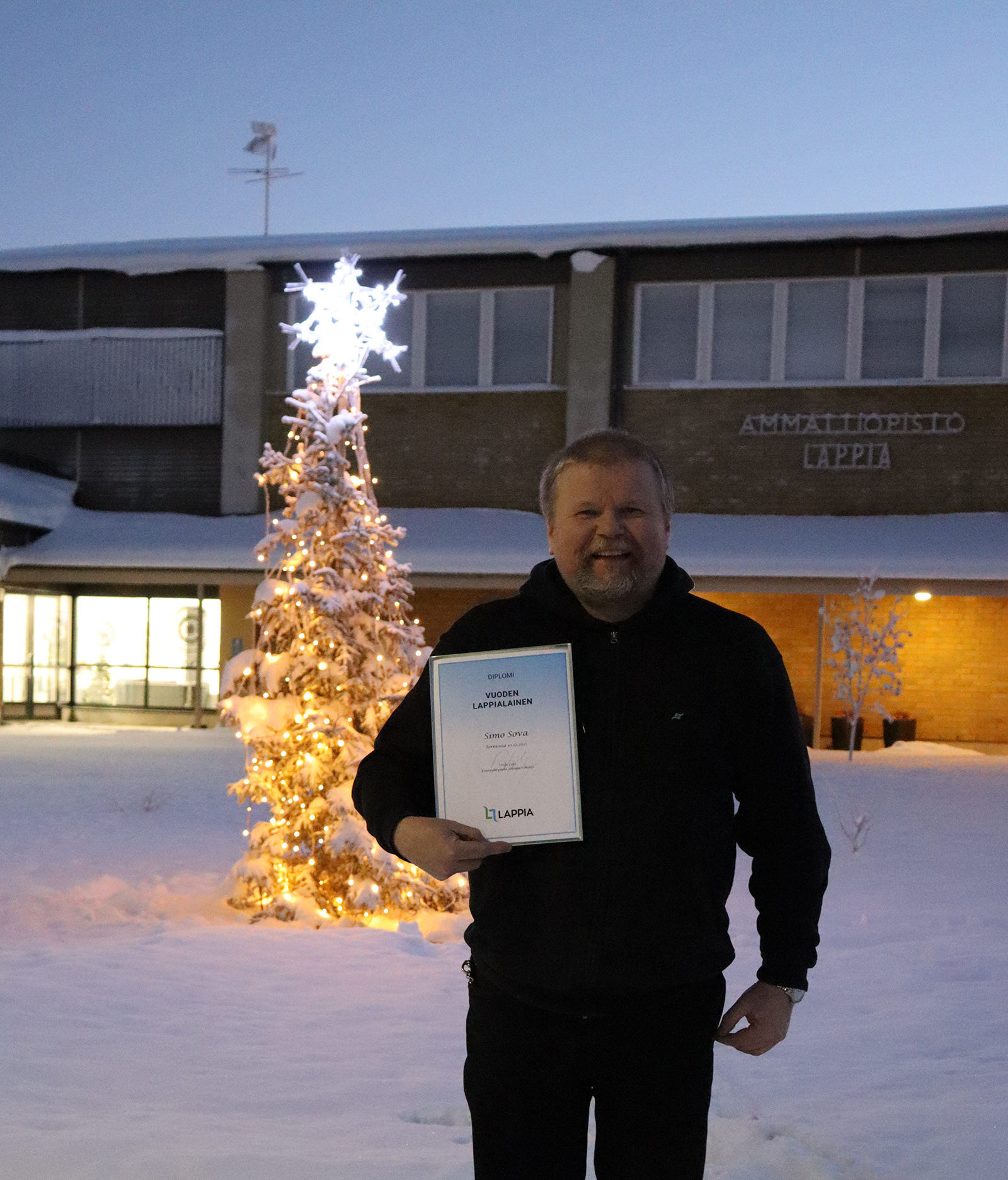 Vuoden lappialainen 2021 seisoo kunniakirja kädessään Muonion toimipaikan lumisella pihalla kunniakirja kädessään. Taustalla on pihalla valaistu joulukuusi.