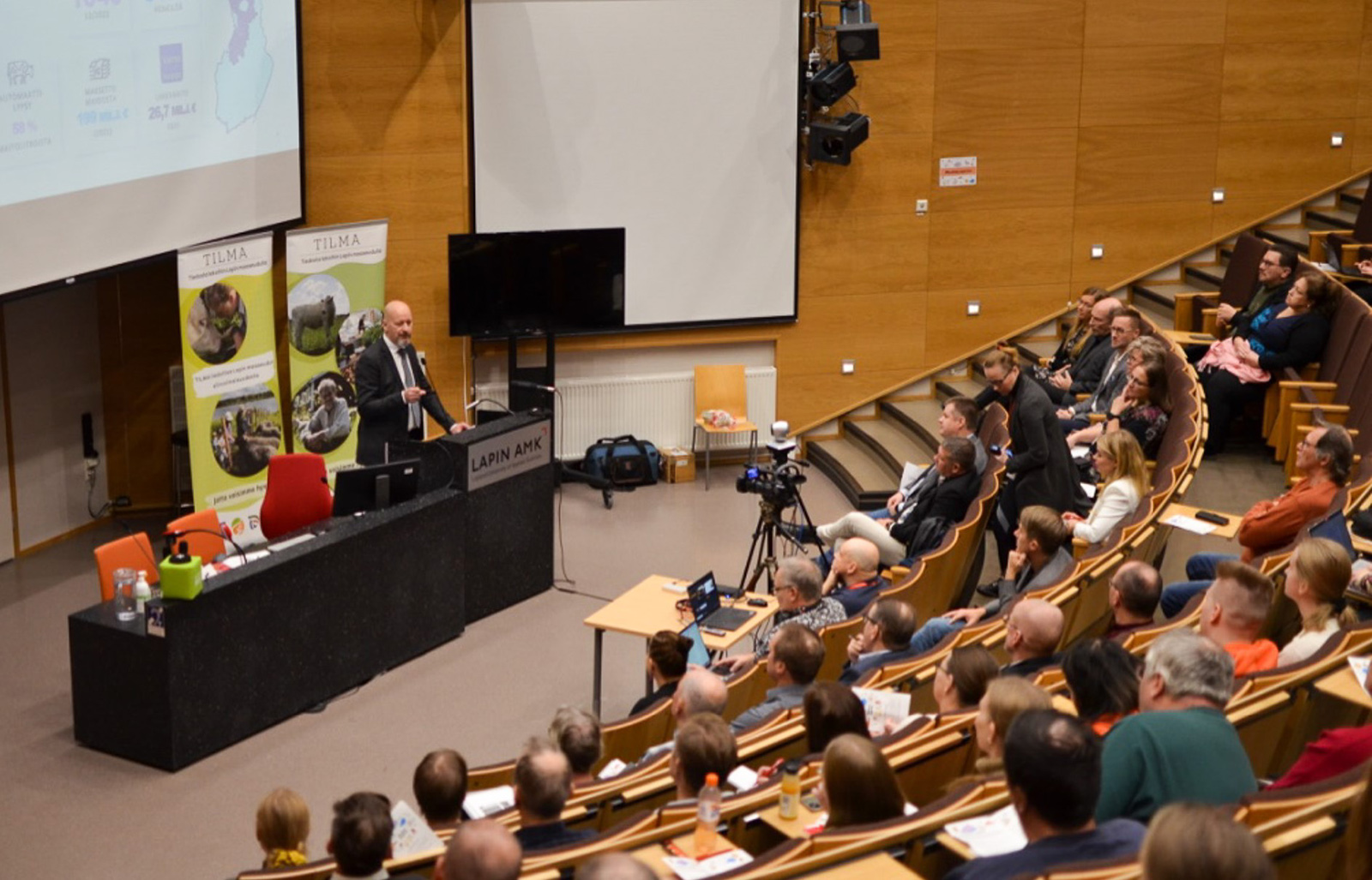 Seminaaritilaisuus auditoriossa, jossa puhuja pöydän takana ja yleisö salissa.