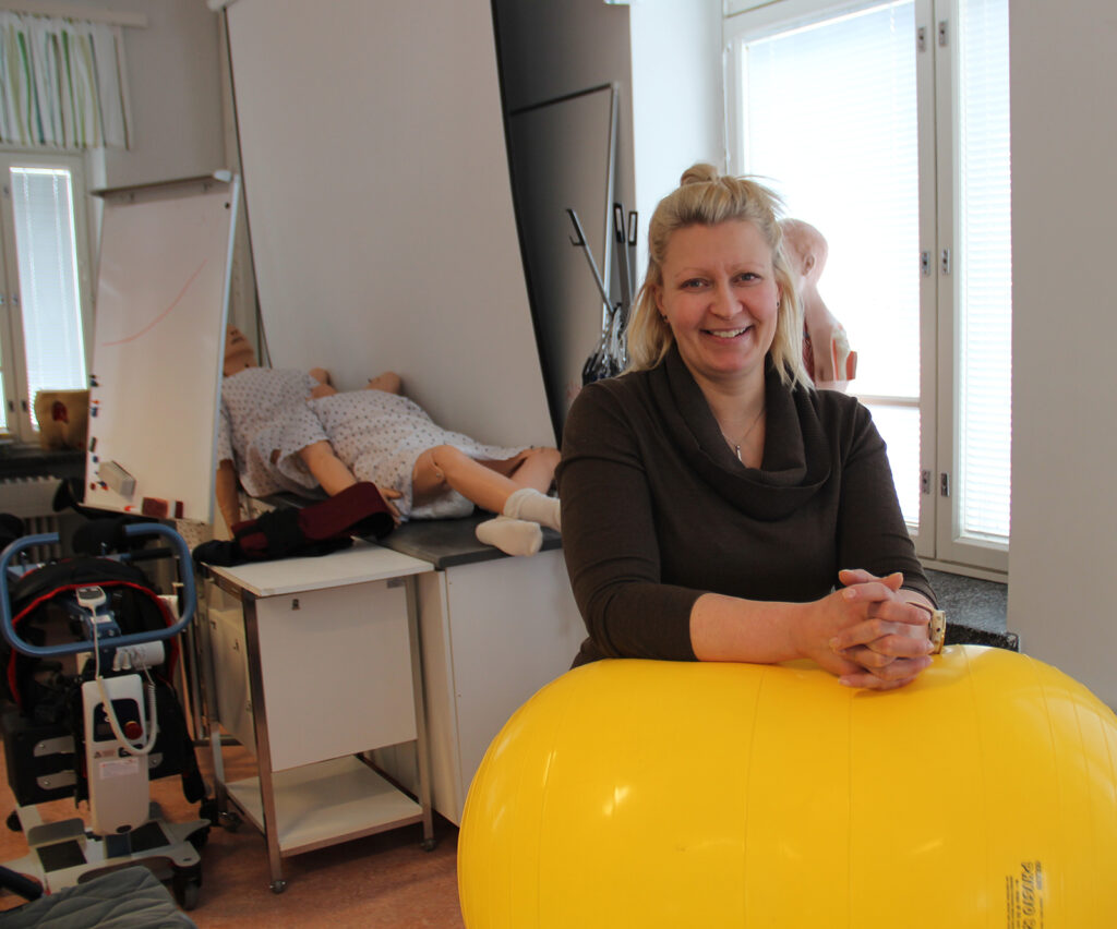 Lähihoitaja Satu Vesa hoitotyön luokassa nojaa  jumppapalloon. Taustalla hoitotyötarvikkeita ja potilasnukkeja.