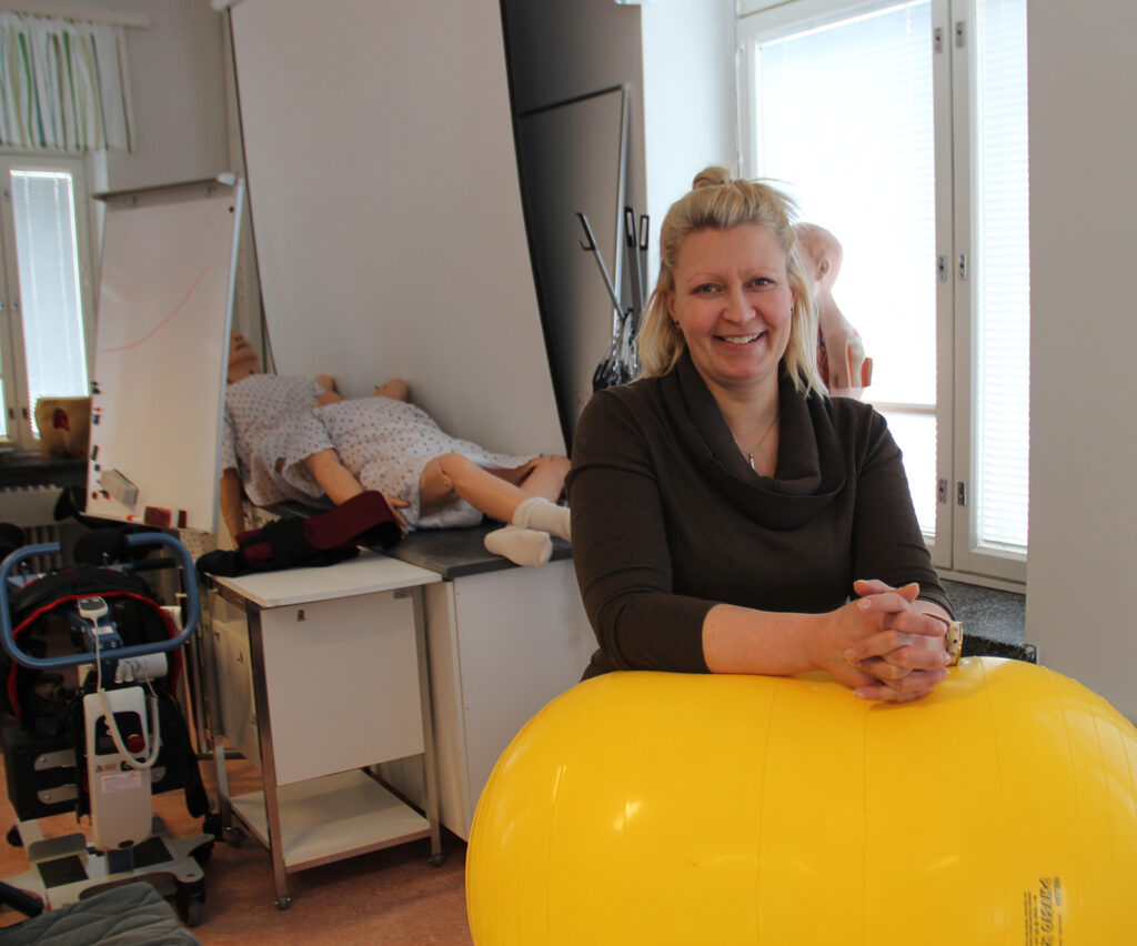 Lähihoitaja Satu Vesa hoitotyön luokassa nojaa jumppapalloon. Taustalla hoitotyötarvikkeita ja potilasnukkeja.