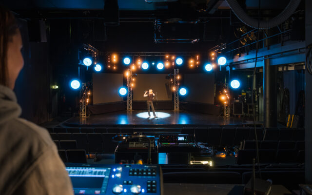 Kuva musiikkitalolta, jossa muusikko on lavalla ja edustalla valopöytä ja sen ohjaaja